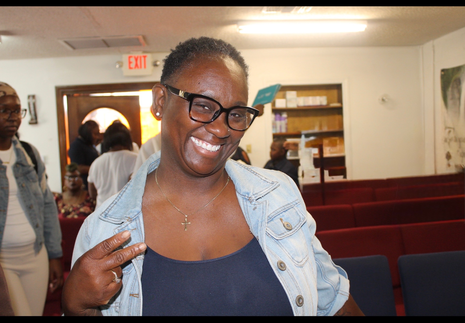 A smiling woman with a peace sign, wearing a cross necklace and denim jacket inside the church